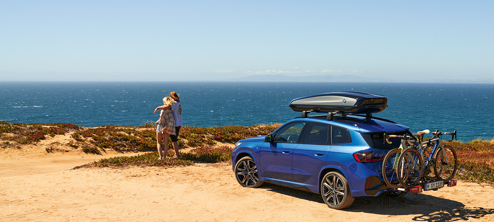 Original BMW Accessories for Spring and Summer BMW X1 U11 standing on sand in front of a coast with 2 people